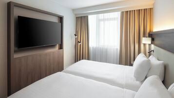 Habitación de hotel con dos camas, un televisor de pantalla plana montado en la pared, una lámpara de noche y una ventana con cortinas.