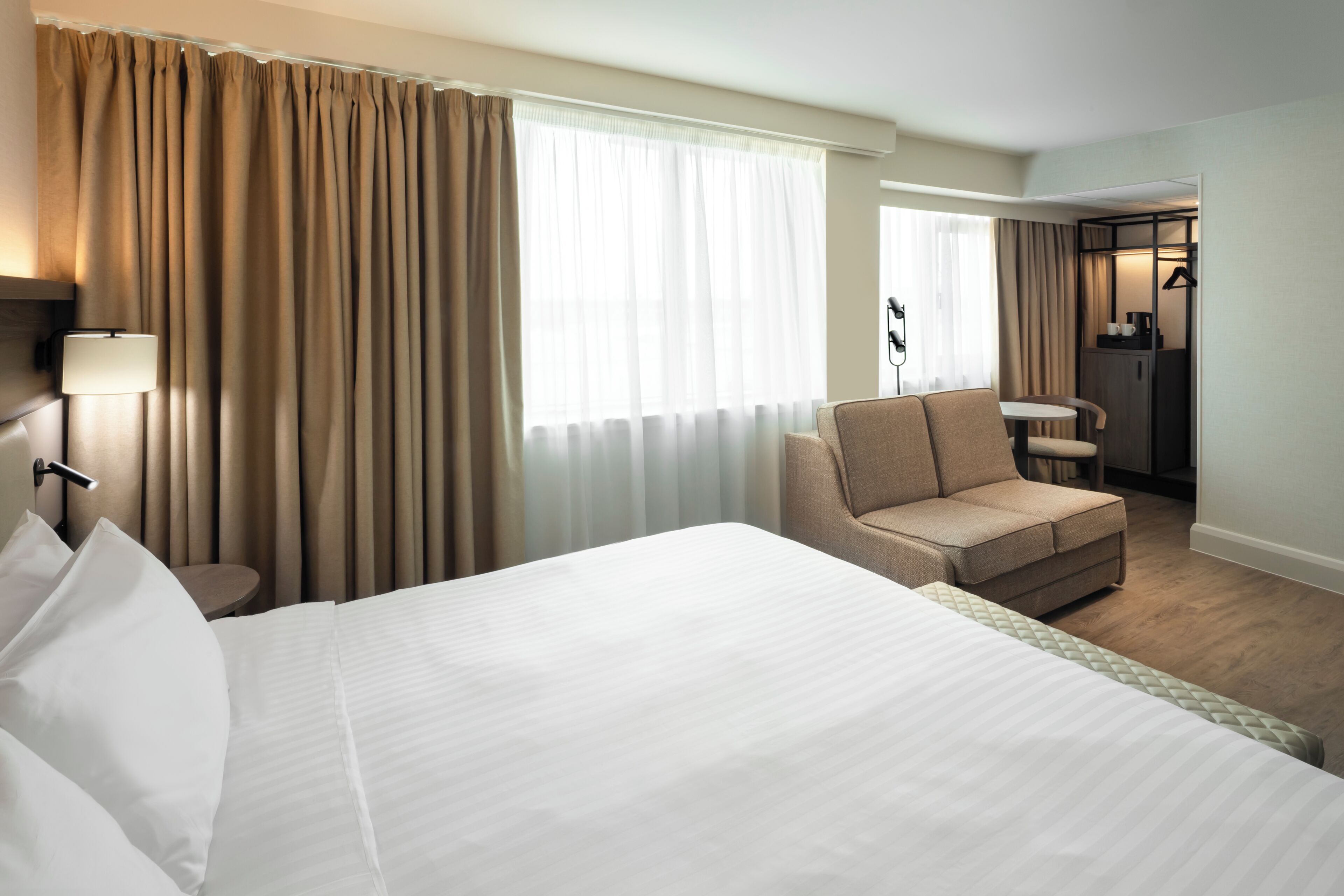 A hotel room with a large bed, a seating area with a chair and table, a desk, and a wardrobe.