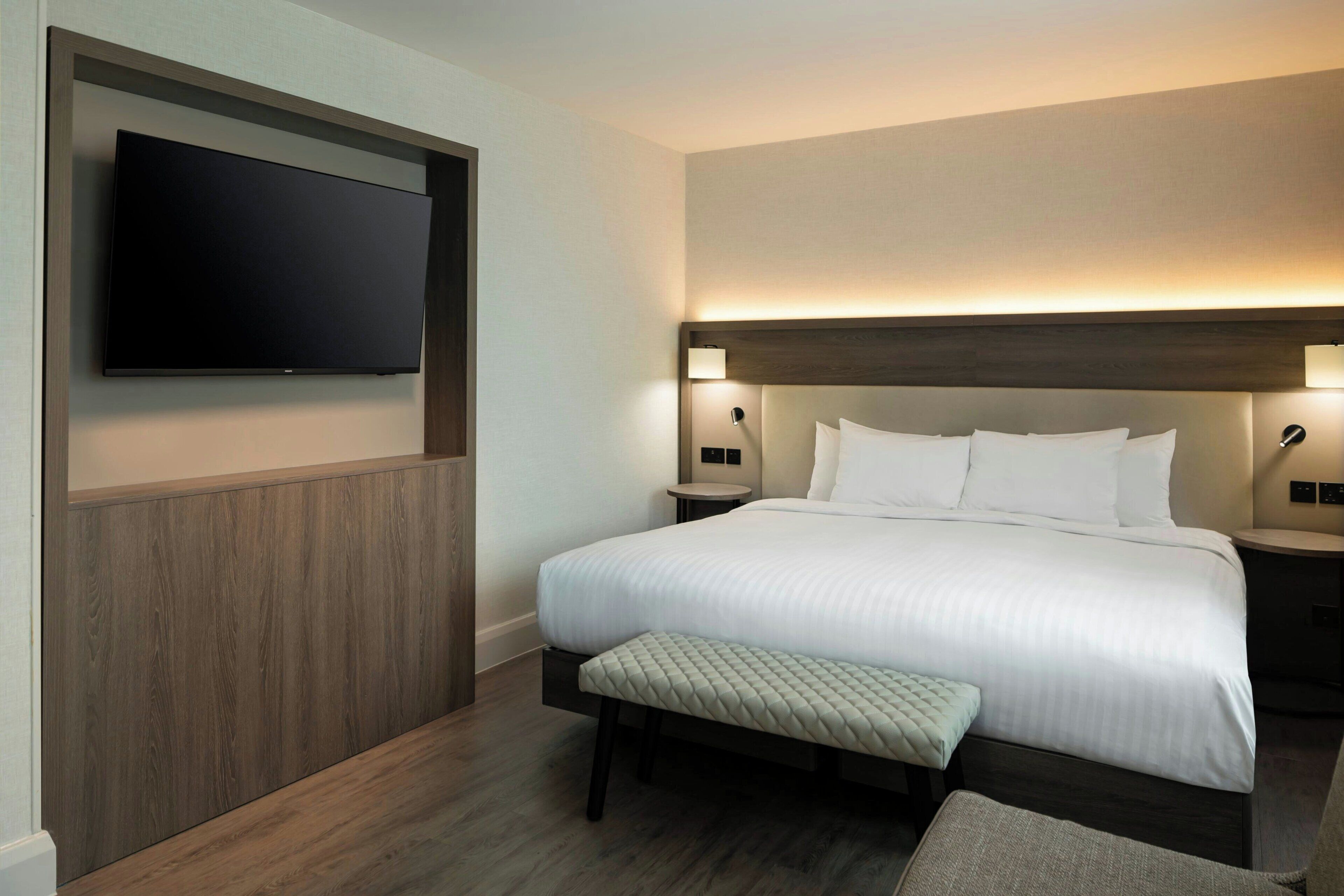 Quarto de hotel com uma cama grande, televisão de tela plana na parede, abajures e detalhes em painéis de madeira.