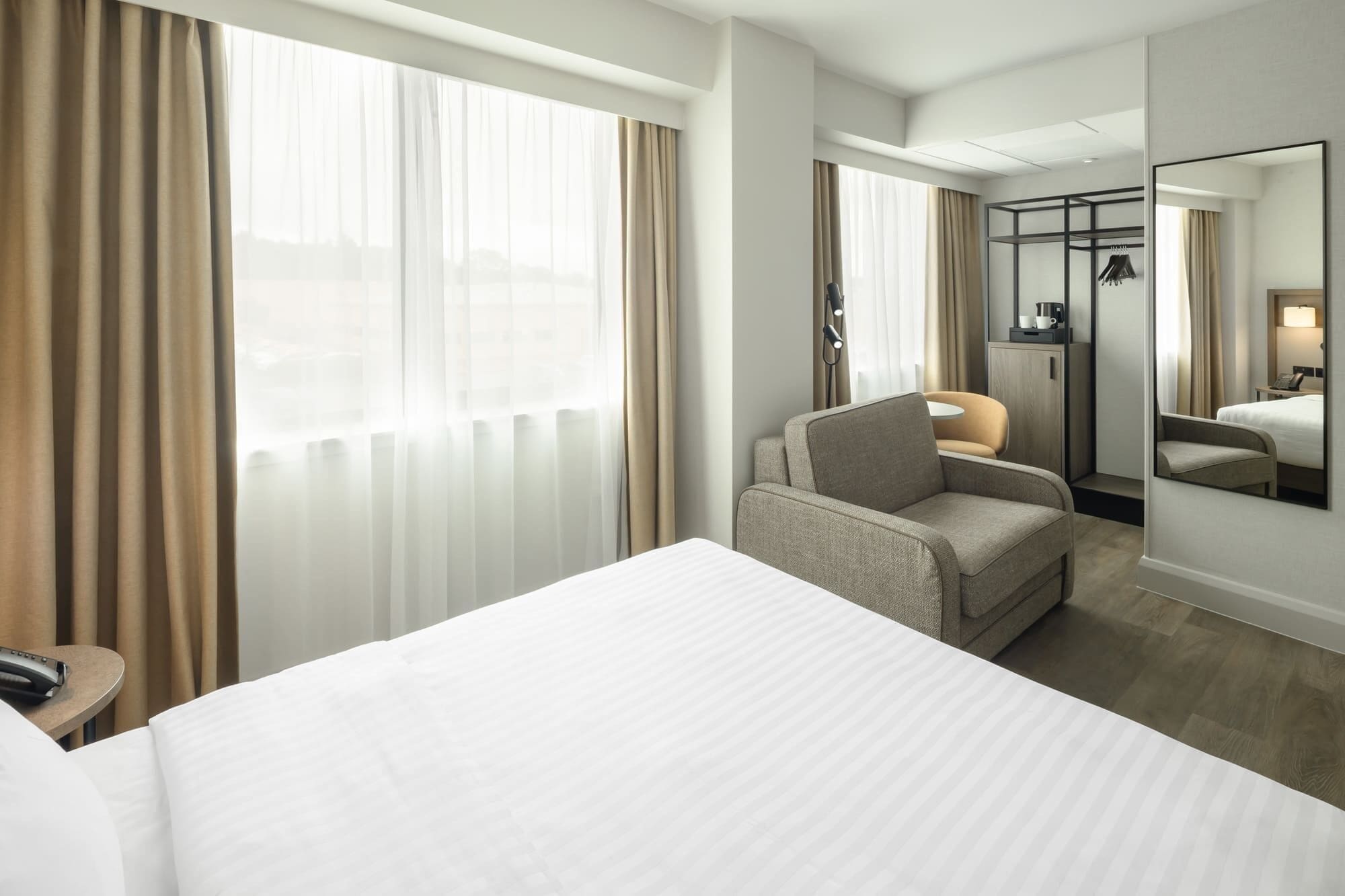A hotel room with a large bed, a chair, a desk, a mirror, and a wardrobe.