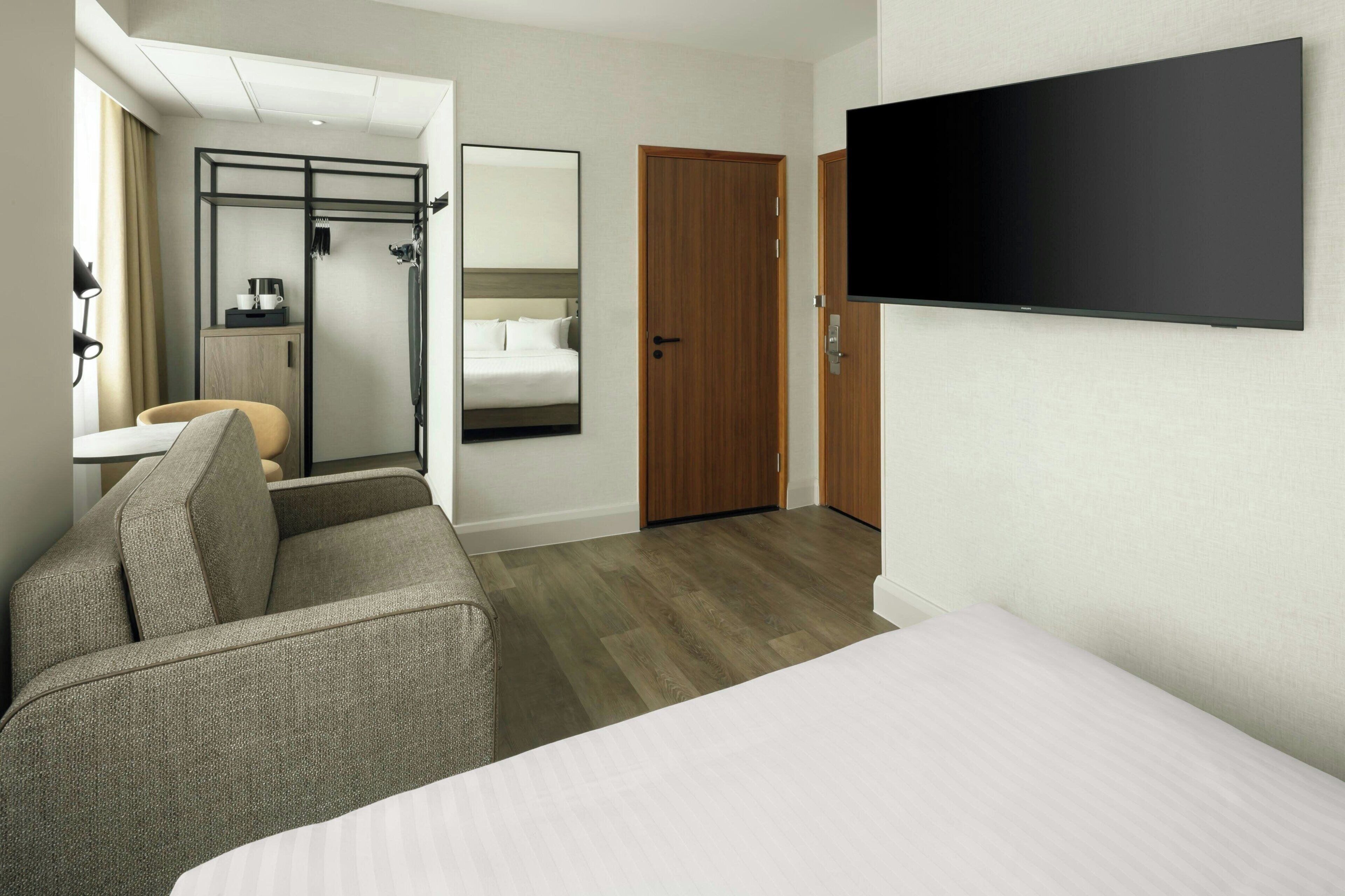 Una habitación de hotel con cama, sofá, televisión, puerta de madera y espejo.