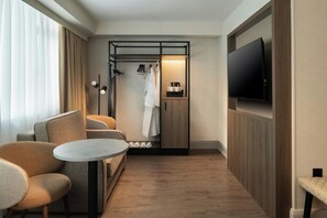 Une chambre d’hôtel compacte comprenant un canapé, une petite table ronde, un téléviseur fixé au mur, une armoire et un espace douche.