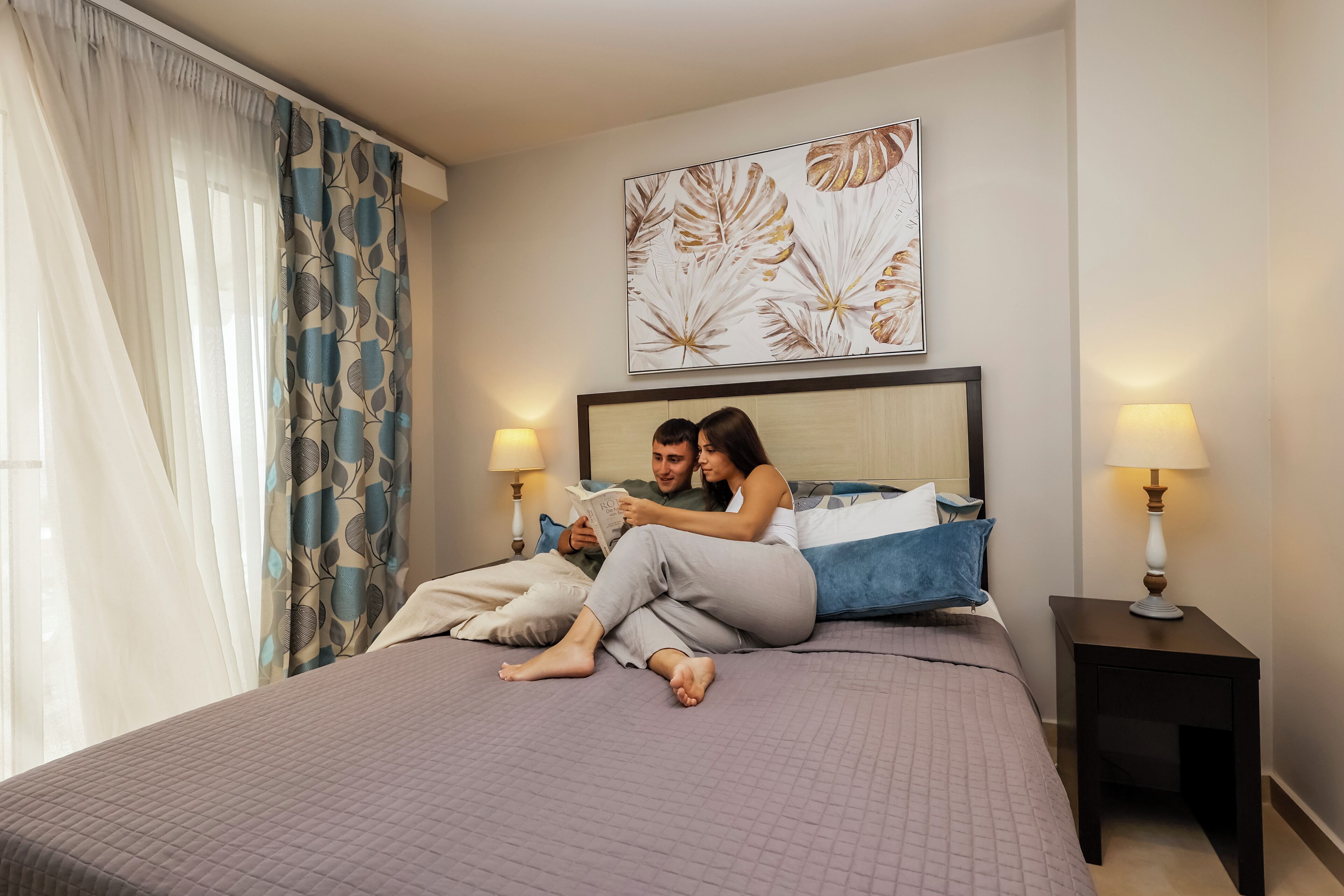 A bedroom with a bed, two bedside lamps, a nightstand, and a window with curtains.