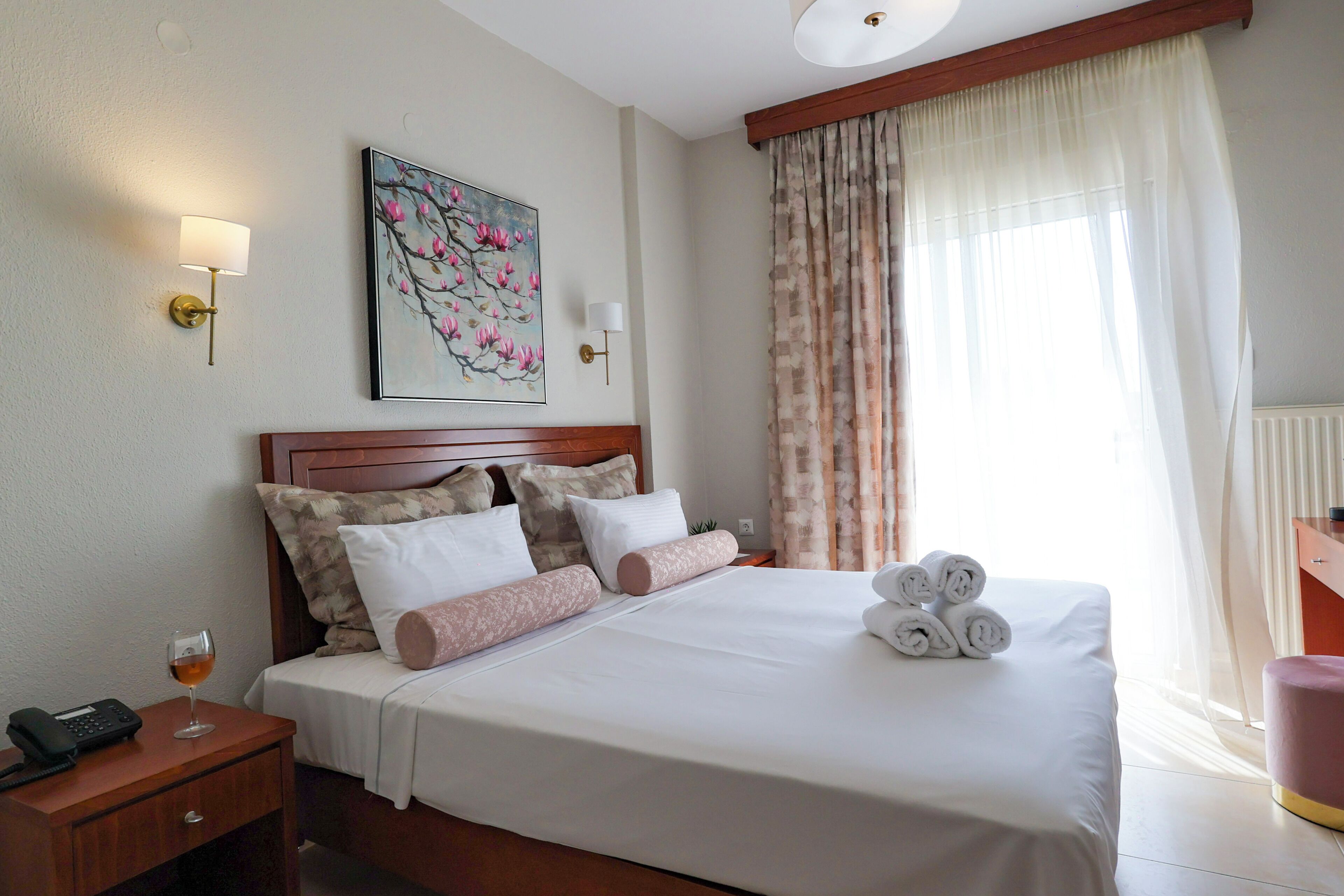 A neatly made bed with white linens and decorative pillows, a bedside table with a lamp and a glass of wine, and a painting of pink flowers on the wall.