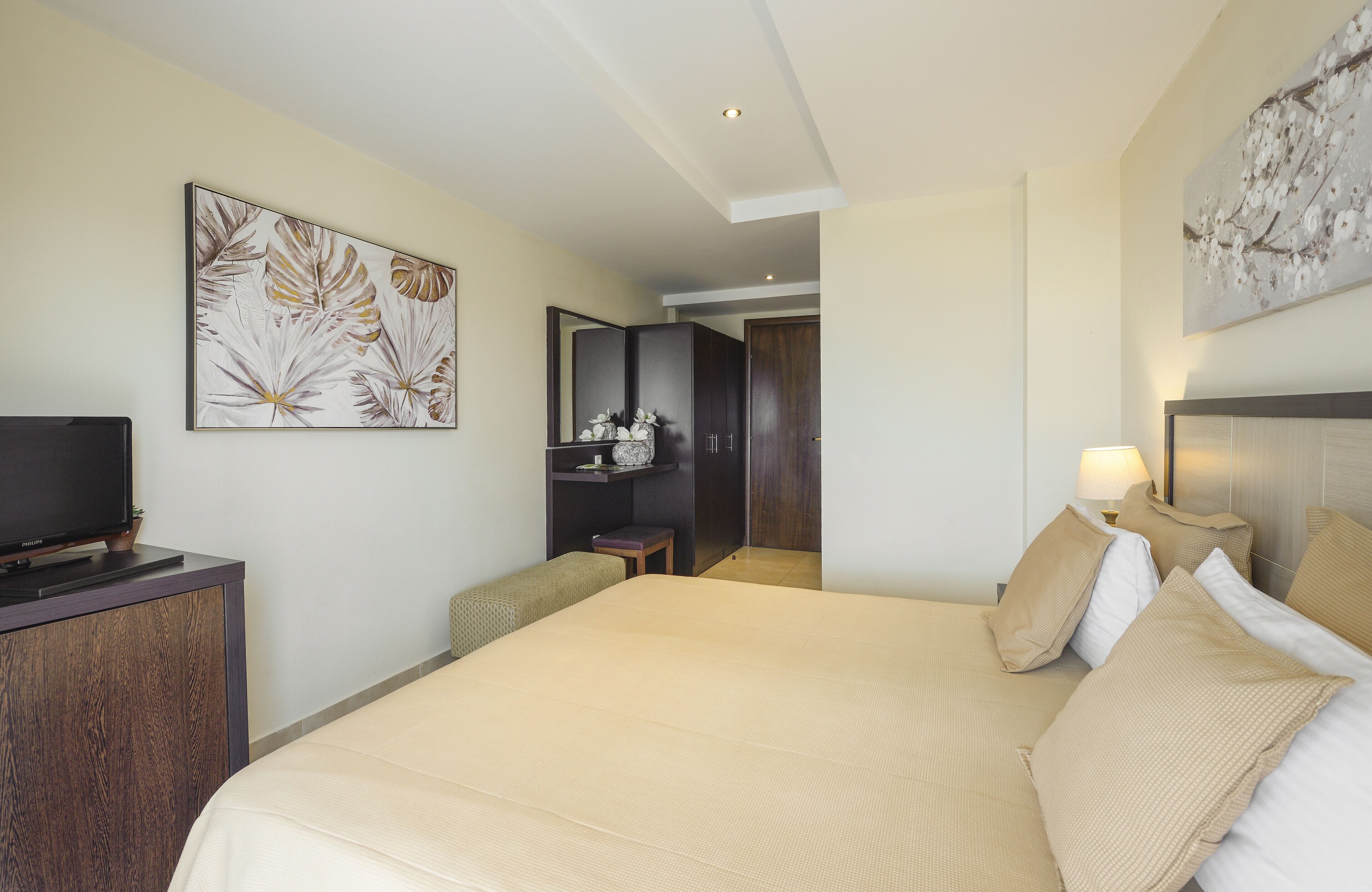A hotel room with a large bed, a television on a wooden cabinet, a desk with a chair, and artwork on the walls.