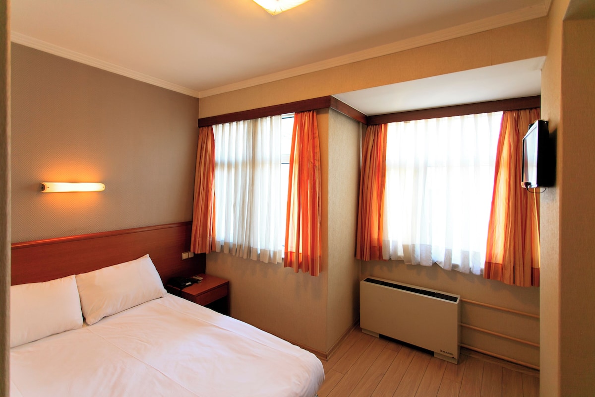 A hotel room with a bed, nightstand, lamp, curtains, and a TV mounted on the wall.