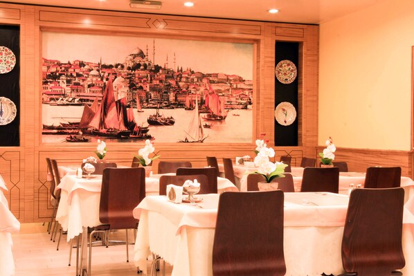 Un comedor con mesas dispuestas para el servicio, un gran mural de un paisaje urbano costero y platos decorativos en las paredes.