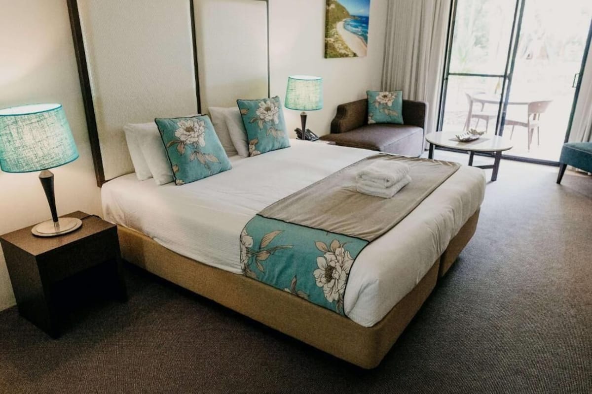 Habitación de hotel con cama, mesita de noche, una silla, una pequeña mesa y vistas al exterior.