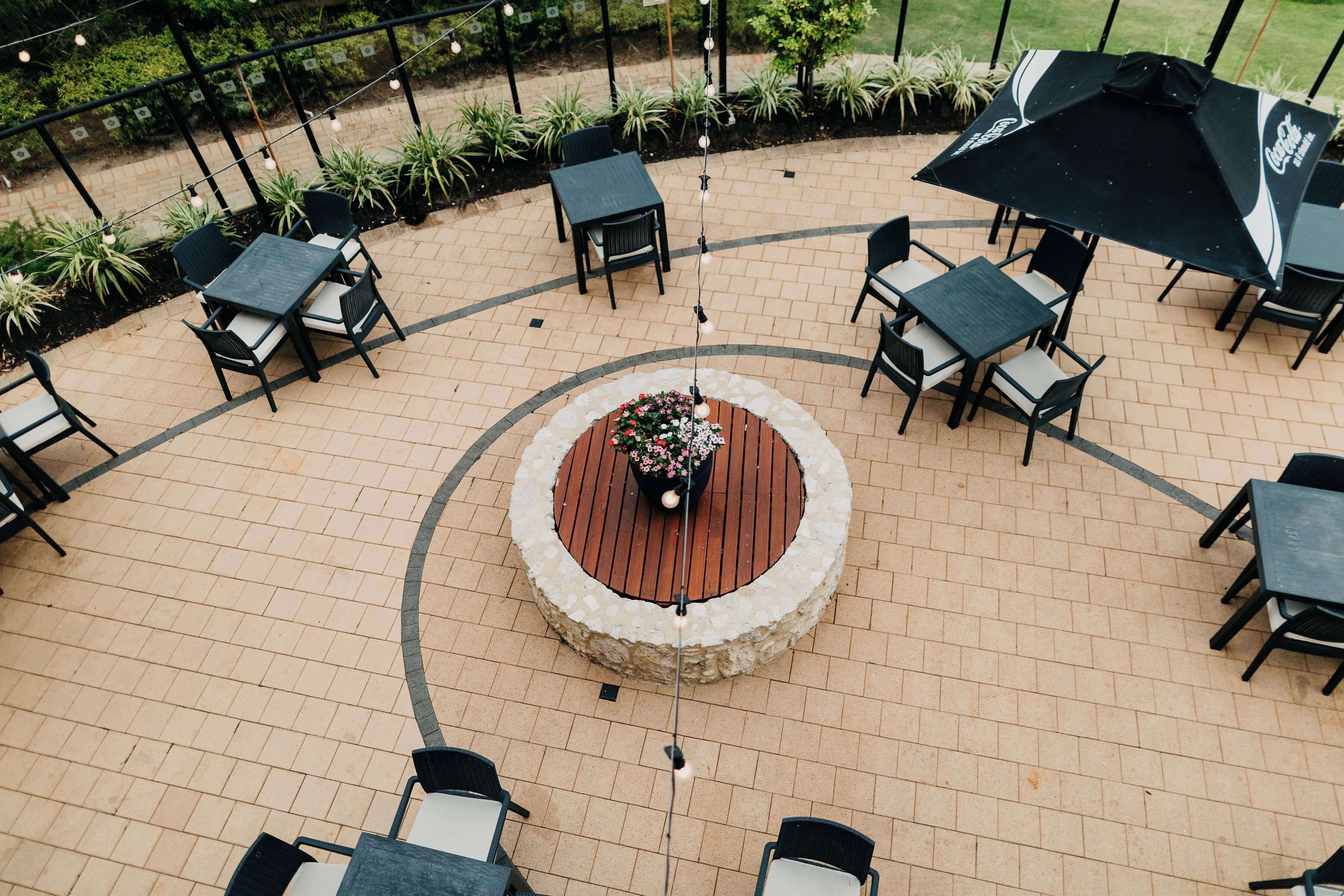 En udendørs patio med runde borde og stole, omgivet af potteplanter og en central træplantekasse med blomster.