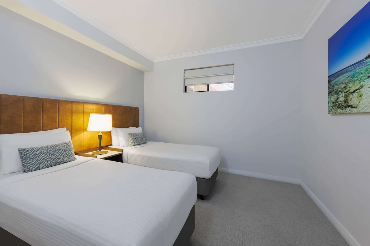 Una habitación de hotel con dos camas, una mesita de noche con lámpara, un ventanal pequeño y un cuadro en la pared.