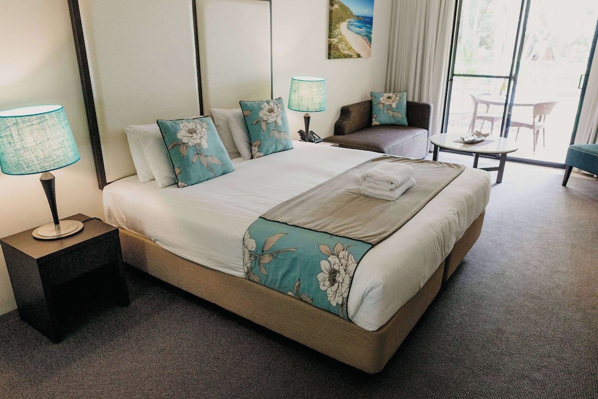 Una cama bien hecha con almohadas florales, una lámpara de noche y vistas al exterior a través de un ventanal grande.