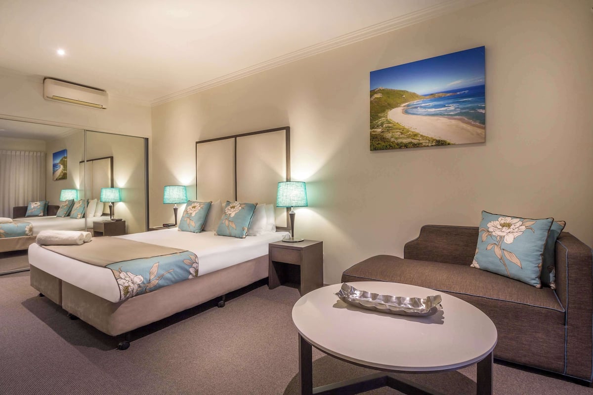 Habitación de hotel con una cama grande, un sofá, una mesa redonda y un cuadro de una playa.