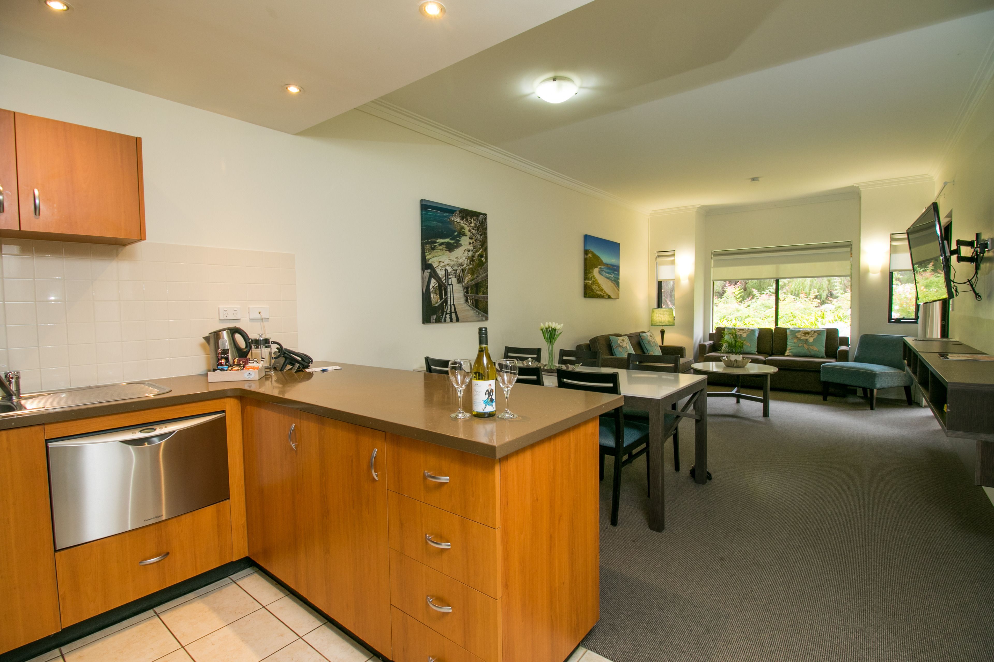 A modern kitchen with wooden cabinets and a built-in dishwasher. There is a dining area with a table and chairs, and a living room with a sofa and a coffee table.