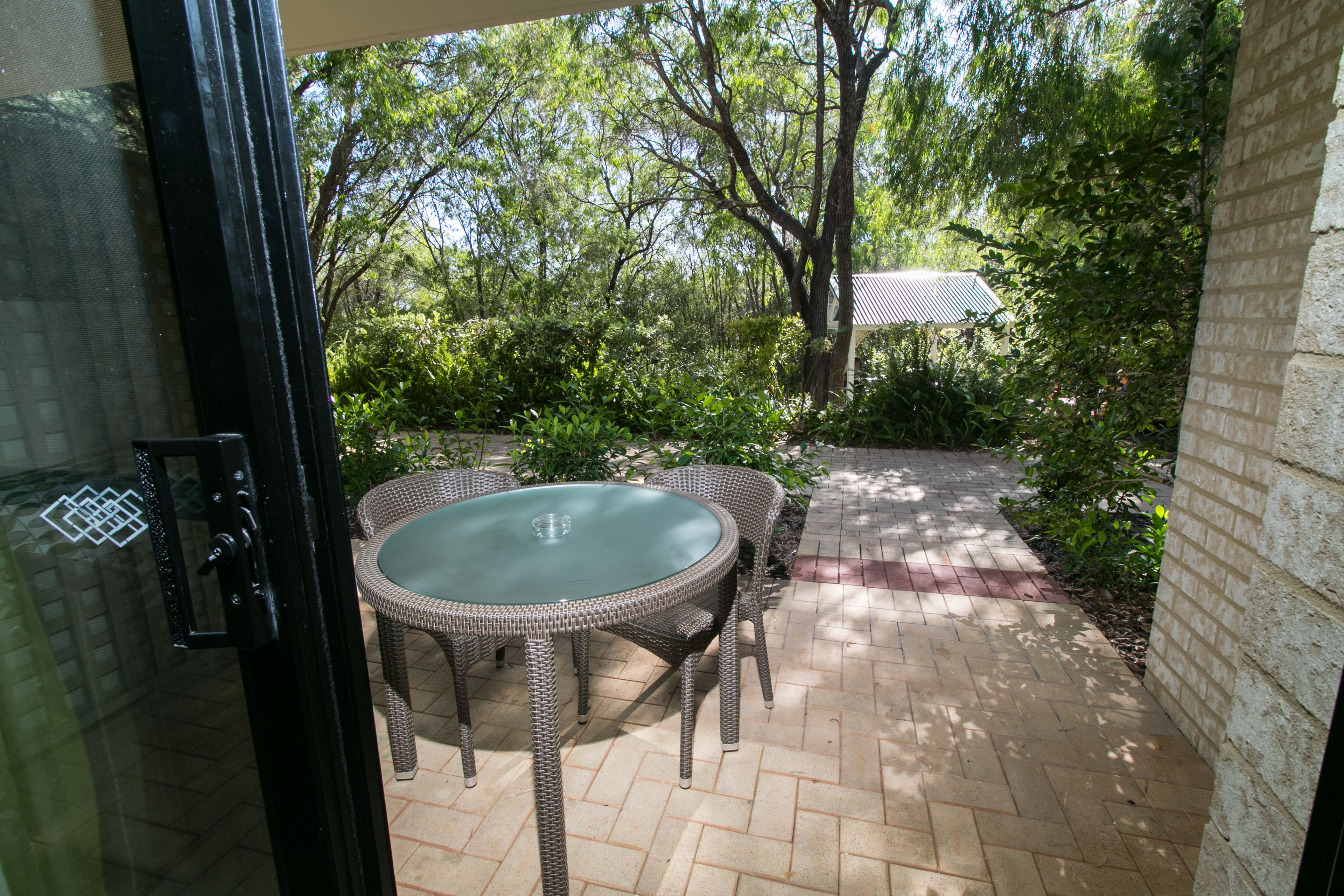 An outdoor patio with wicker furniture and a glass table, surrounded by greenery and a brick pathway.