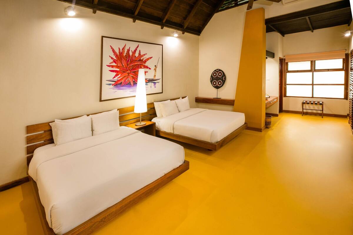 Quarto de hotel com duas camas, cabeceira de madeira, obra de arte na parede, janela com persianas e um pequeno banco.