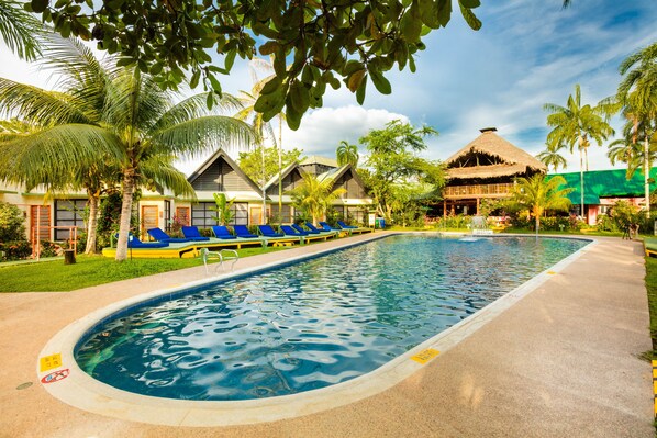 A swimming pool with lounge chairs, palm trees, and a thatched-roof building. - Decameron Decalodge Ticuna (Leticia)