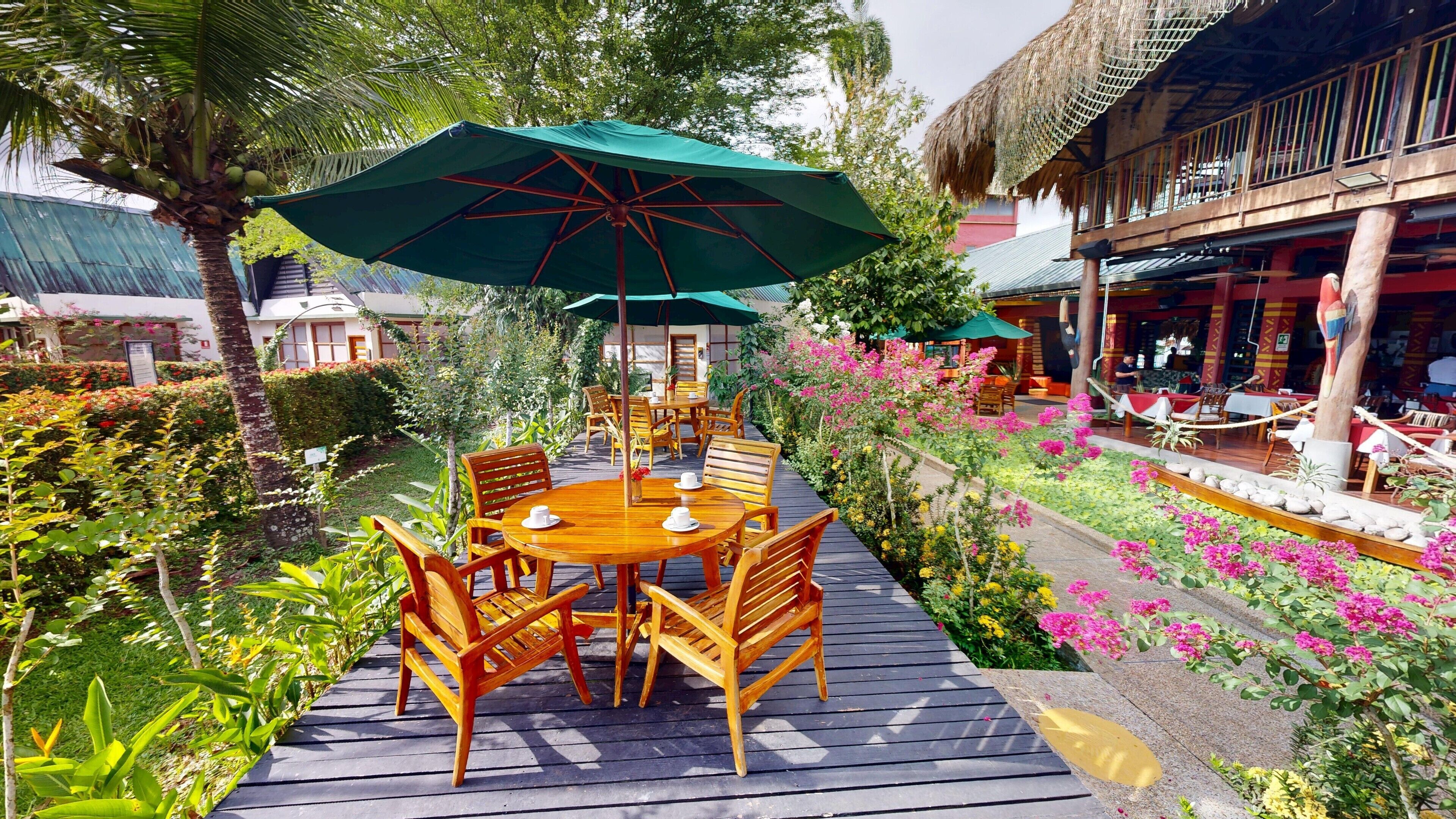 Un área de comedor al aire libre con mesas y sillas de madera, rodeada de abundante vegetación y plantas con flores.