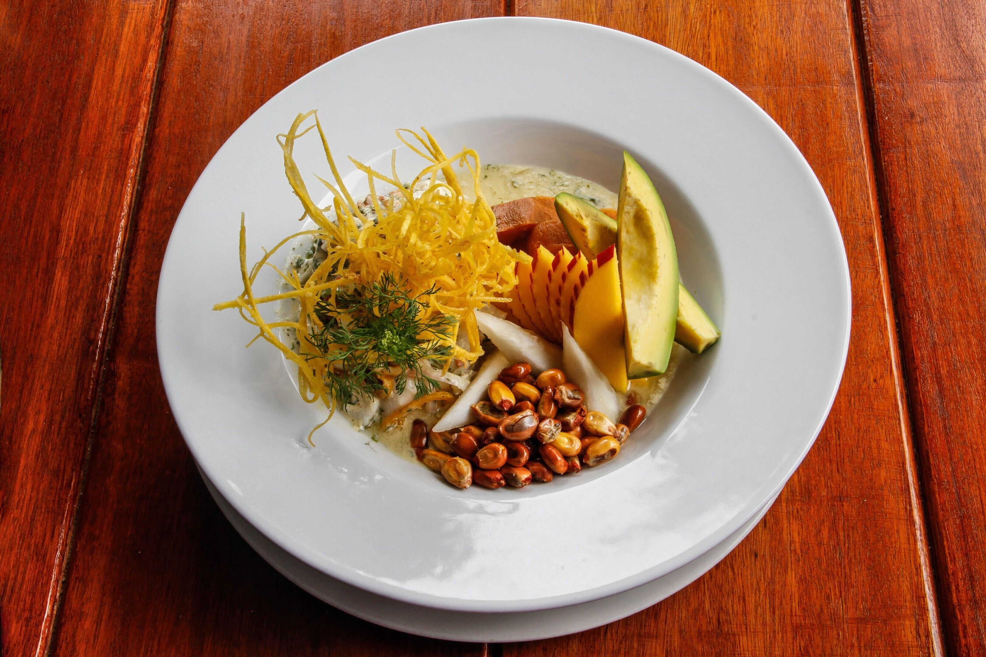 Een wit bord met diverse soorten eten, waaronder avocado, noten en een romige saus, op een houten tafel.