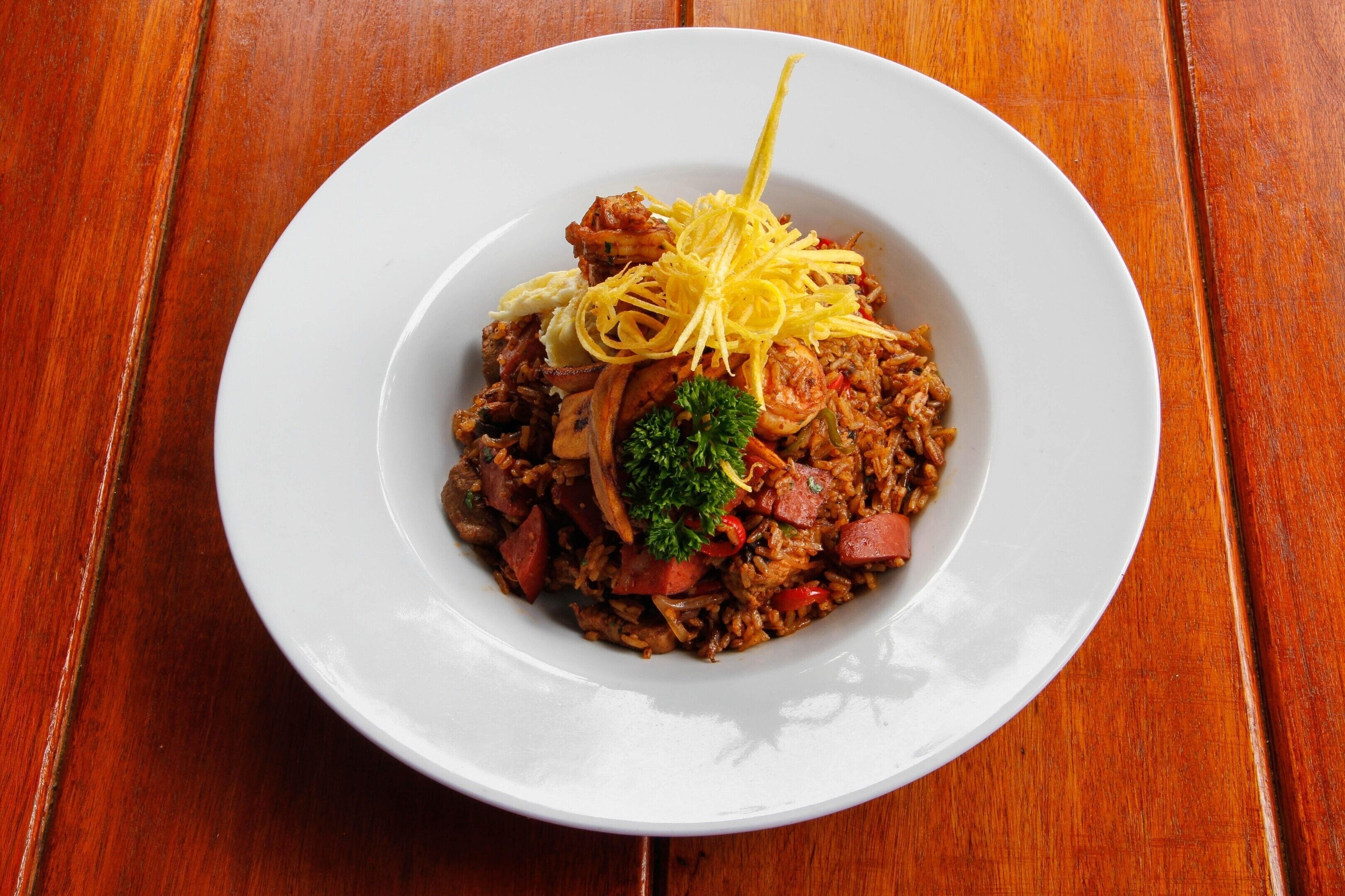Une assiette blanche contenant un plat de riz, de saucisses et de légumes, garni de persil et de zeste de citron, posée sur une table en bois.
