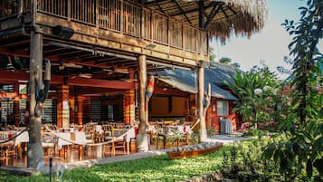 Un restaurant a l'aire lliure amb taules i cadires de fusta, un teulat de palla i una zona de jardí.