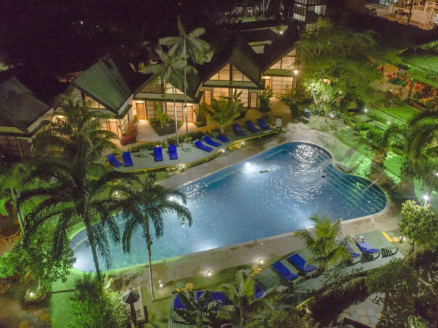 Una piscina rodeada de sillas de descanso y palmeras, con un edificio al fondo.
