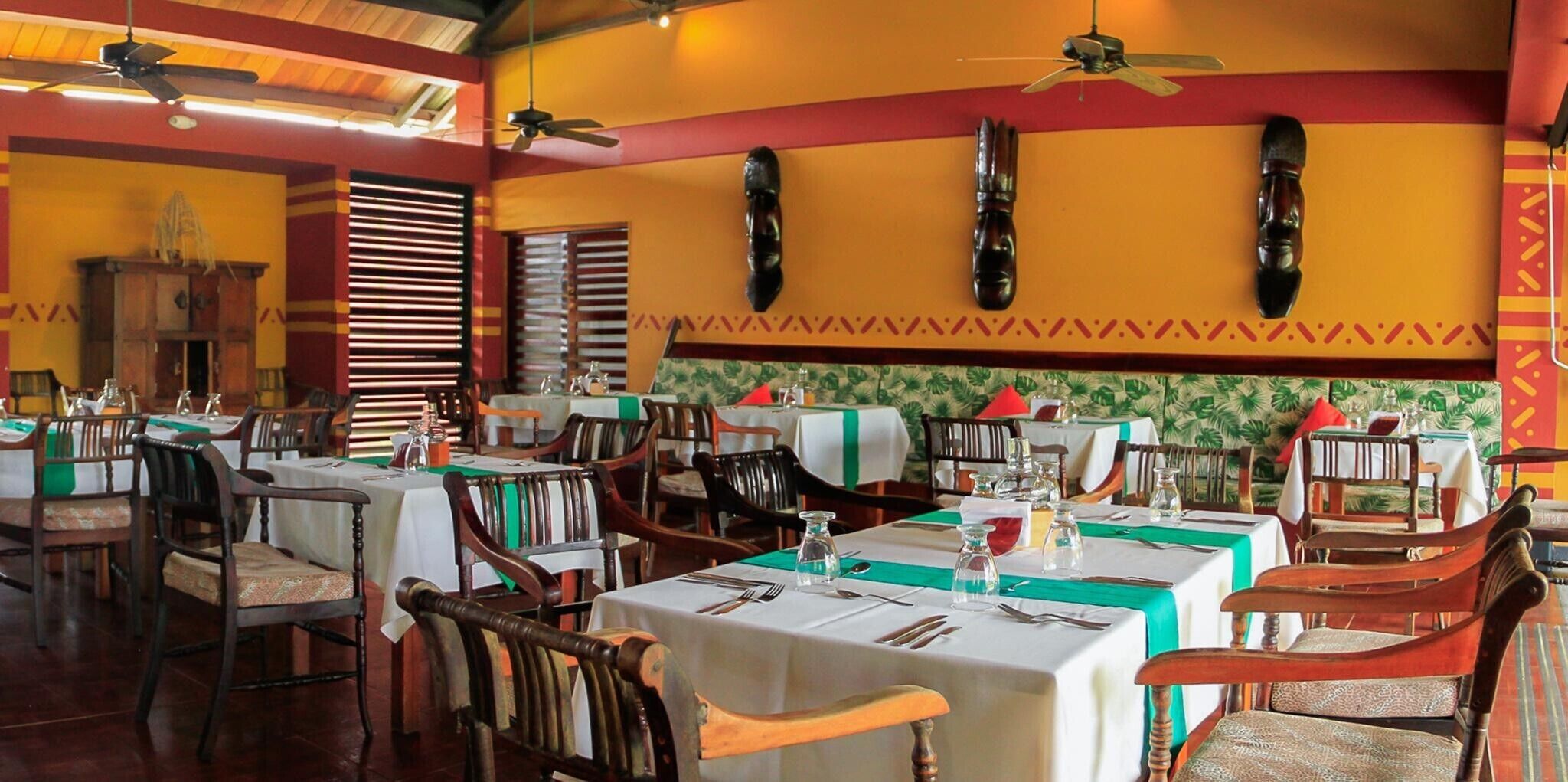 A restaurant with multiple tables, chairs, ceiling fans, and a tropical wallpaper design.