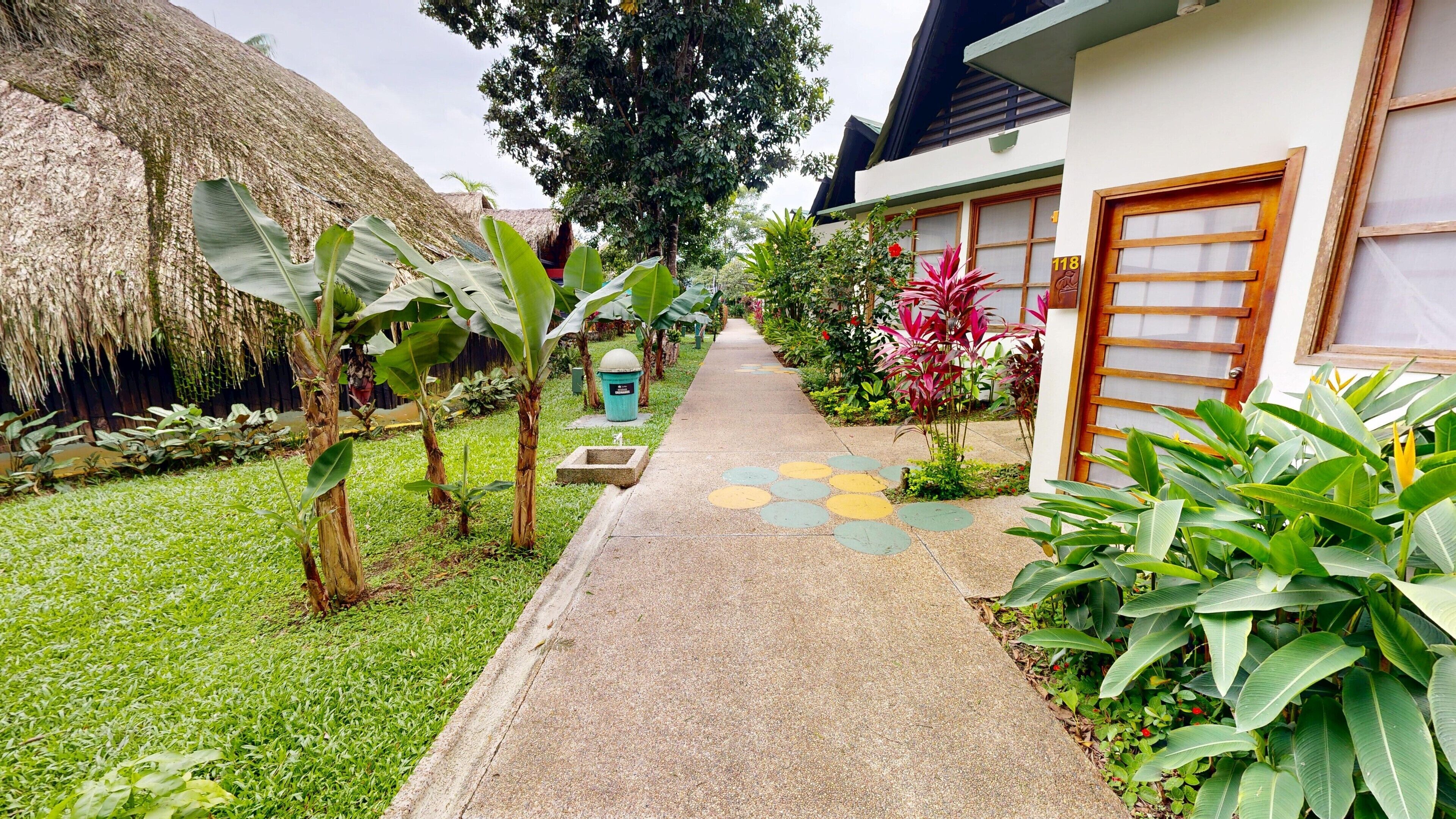 A pathway leading to a building with a number 118 sign, surrounded by lush greenery and a thatched structure.