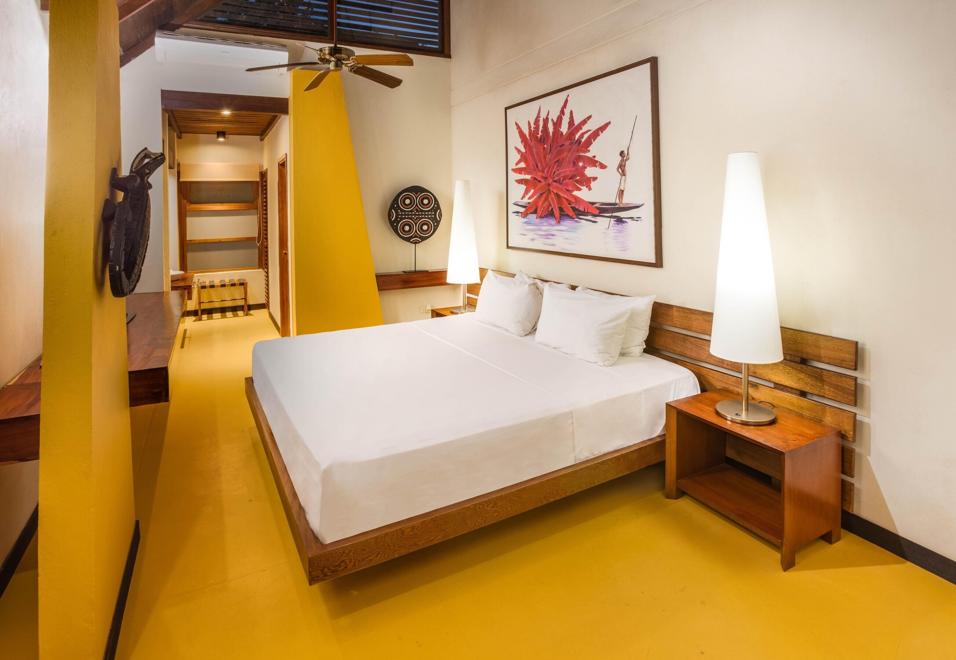 Habitación de hotel con una cama, dos lámparas de noche, un ventilador de techo, un cuadro colgado en la pared y un banco de madera.