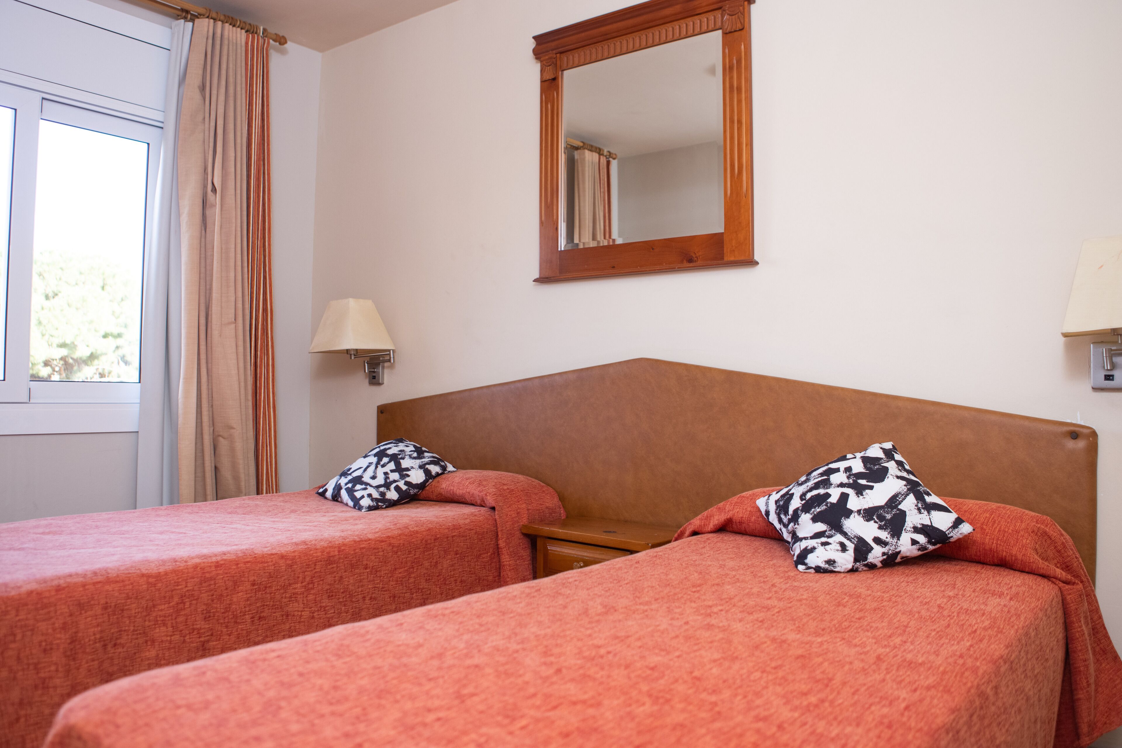 A hotel room with two single beds, each with a patterned pillow, a wooden headboard, a mirror, and a window with curtains.