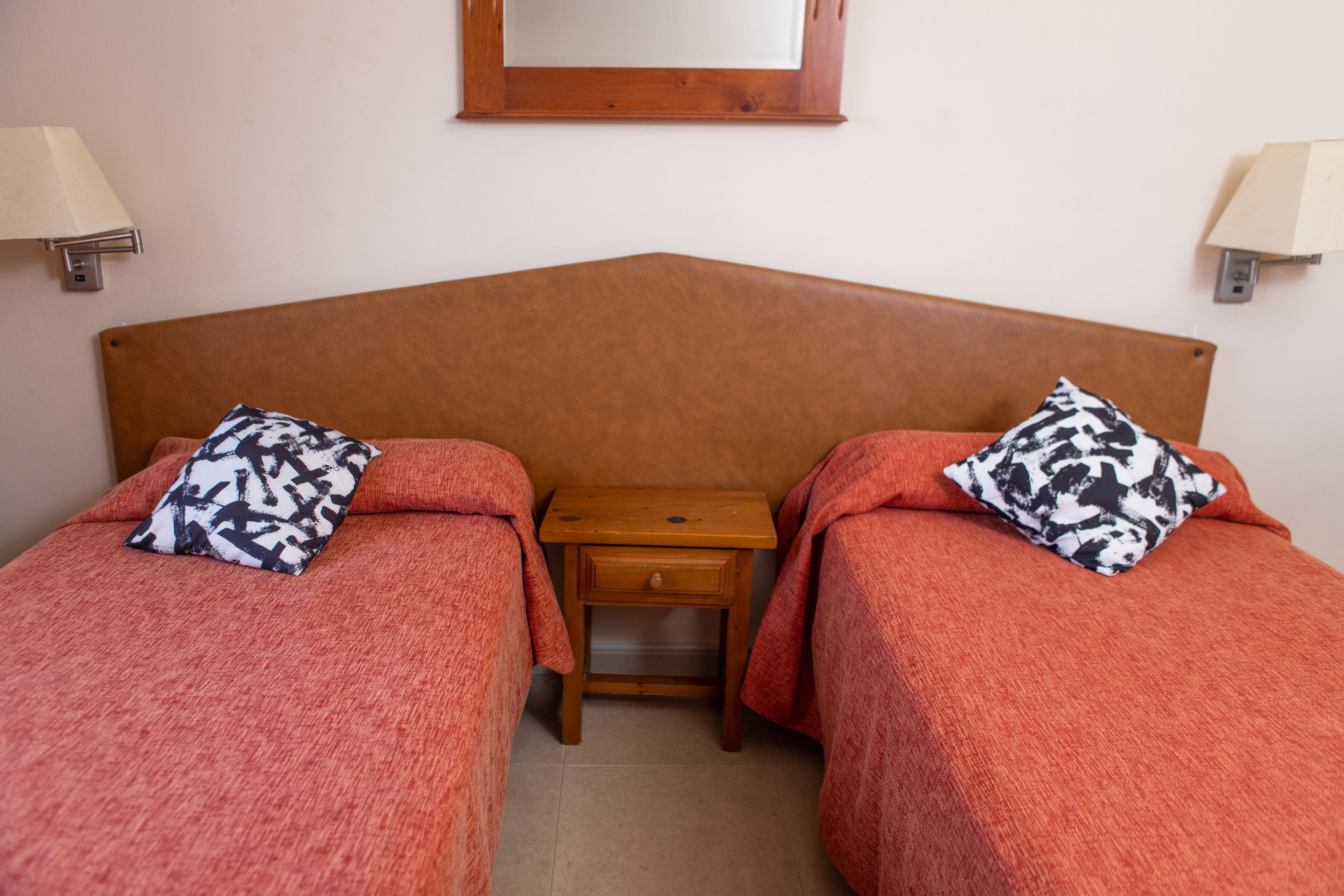 Una habitación con dos camas, cada una con una almohada estampada, una mesita de noche de madera y un espejo en la pared.