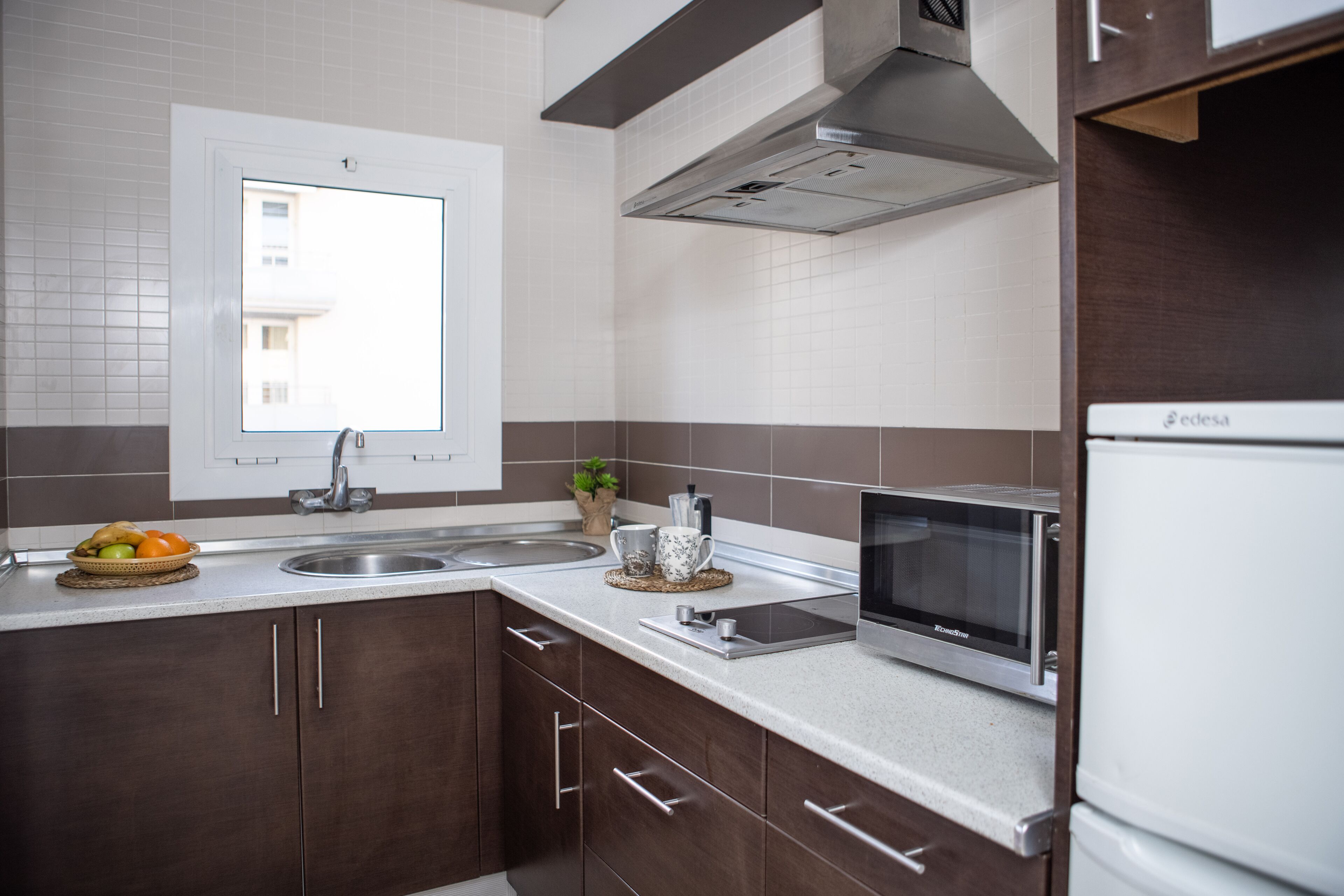 A modern kitchen with a stainless steel sink, a microwave, and a built-in oven. There is a window above the sink and a potted plant on the counter.