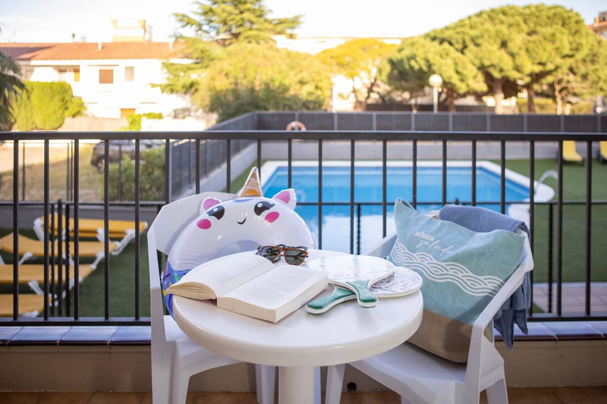A balcony with a view of a swimming pool, a white chair, a book, a pair of sunglasses, and a beach-themed pillow.
