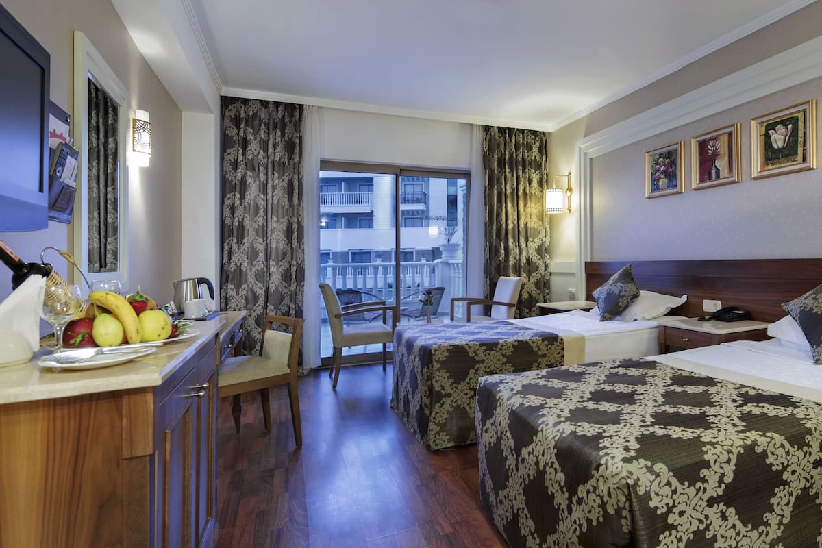Habitación de hotel con dos camas, un escritorio con fruta y una botella, una silla, vistas al balcón y un televisor.