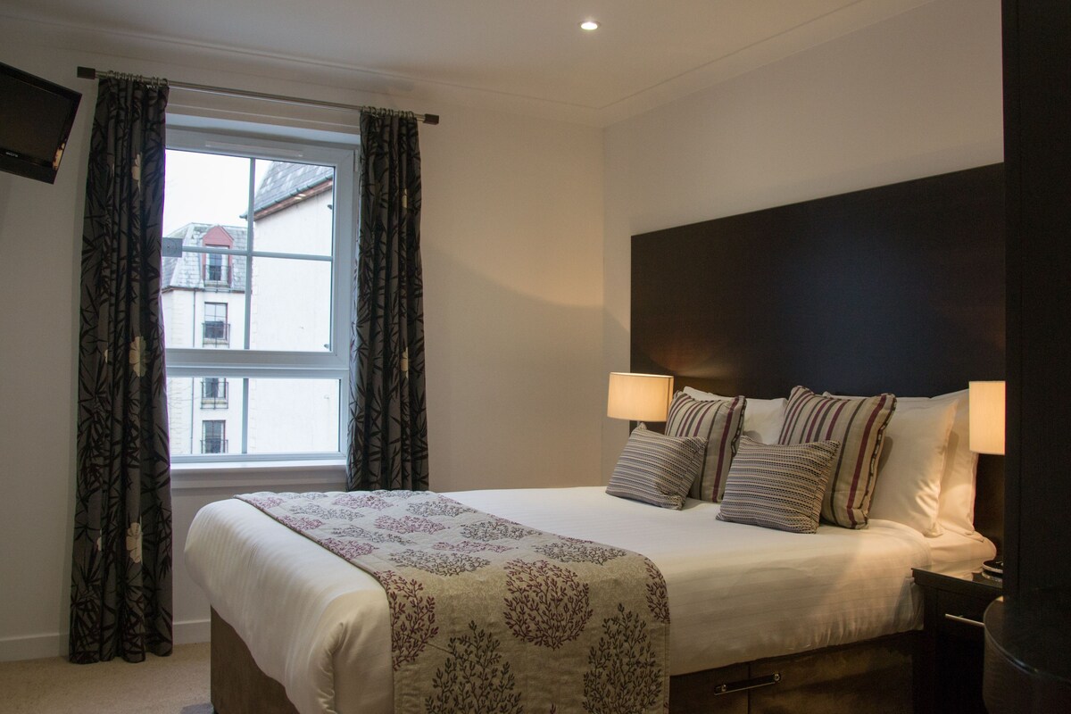 A neatly made bed with a patterned bedspread, two bedside lamps, and a dark headboard.