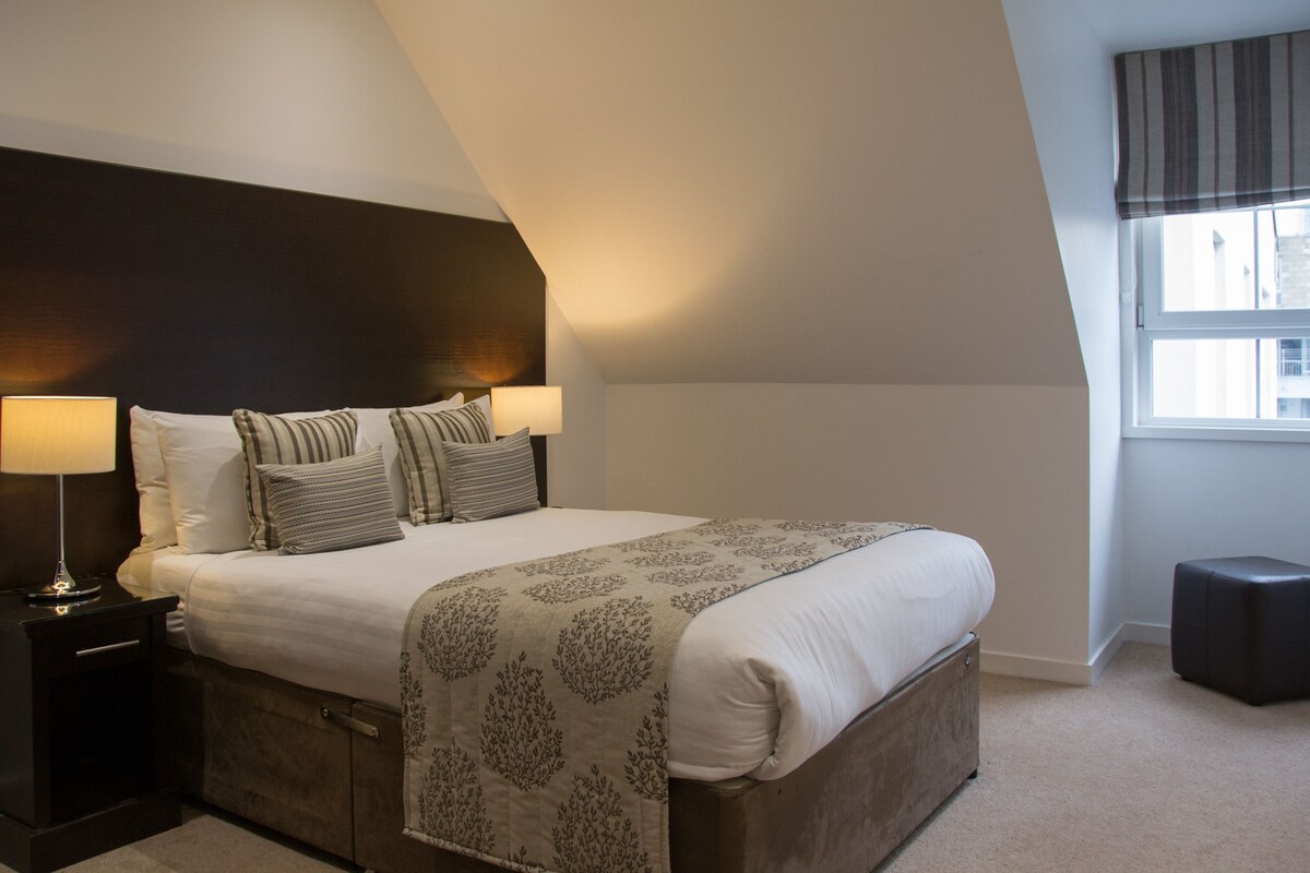 A bedroom with a large bed, two bedside lamps, a dark headboard, a window with a view, and a small ottoman.