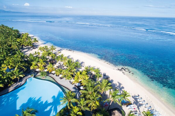 Vista aérea de un complejo turístico con piscina, palmeras y un océano azul cristalino.