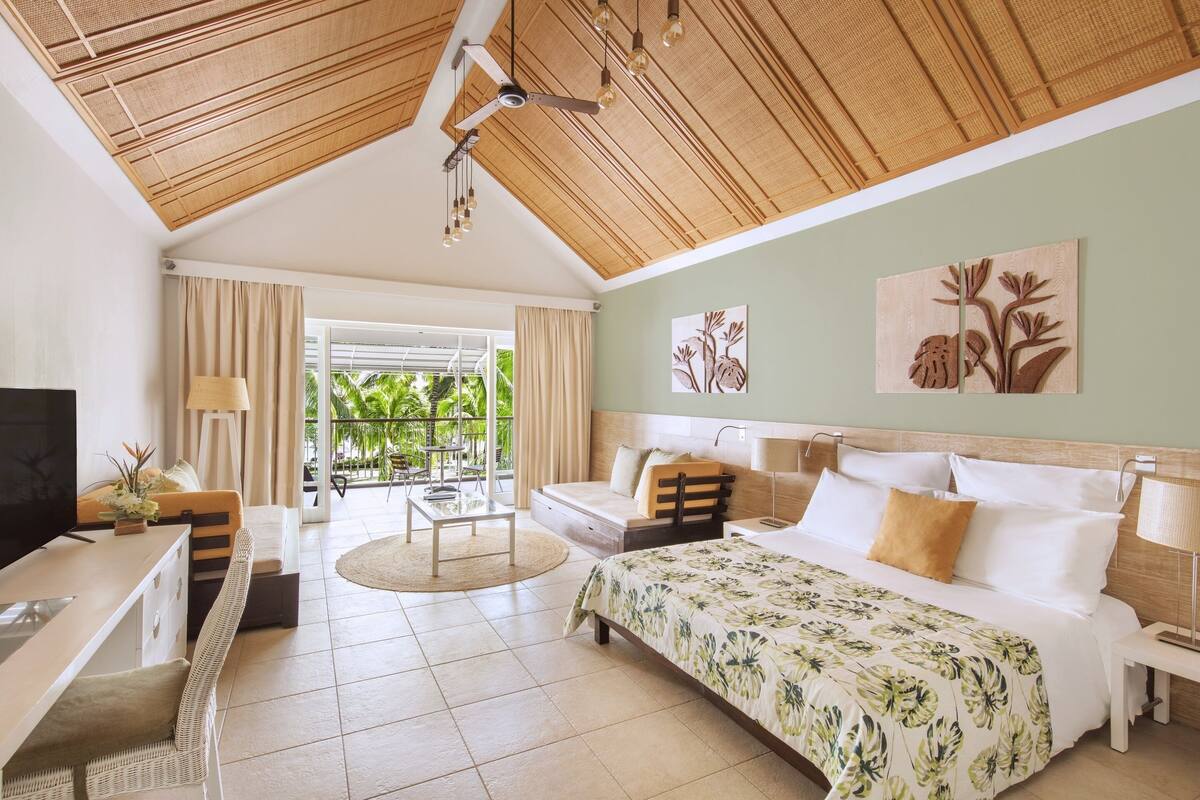 Habitación amplia con cama, escritorio y zona de estar. Se aprecia una ventana con cortinas y vistas al exterior.