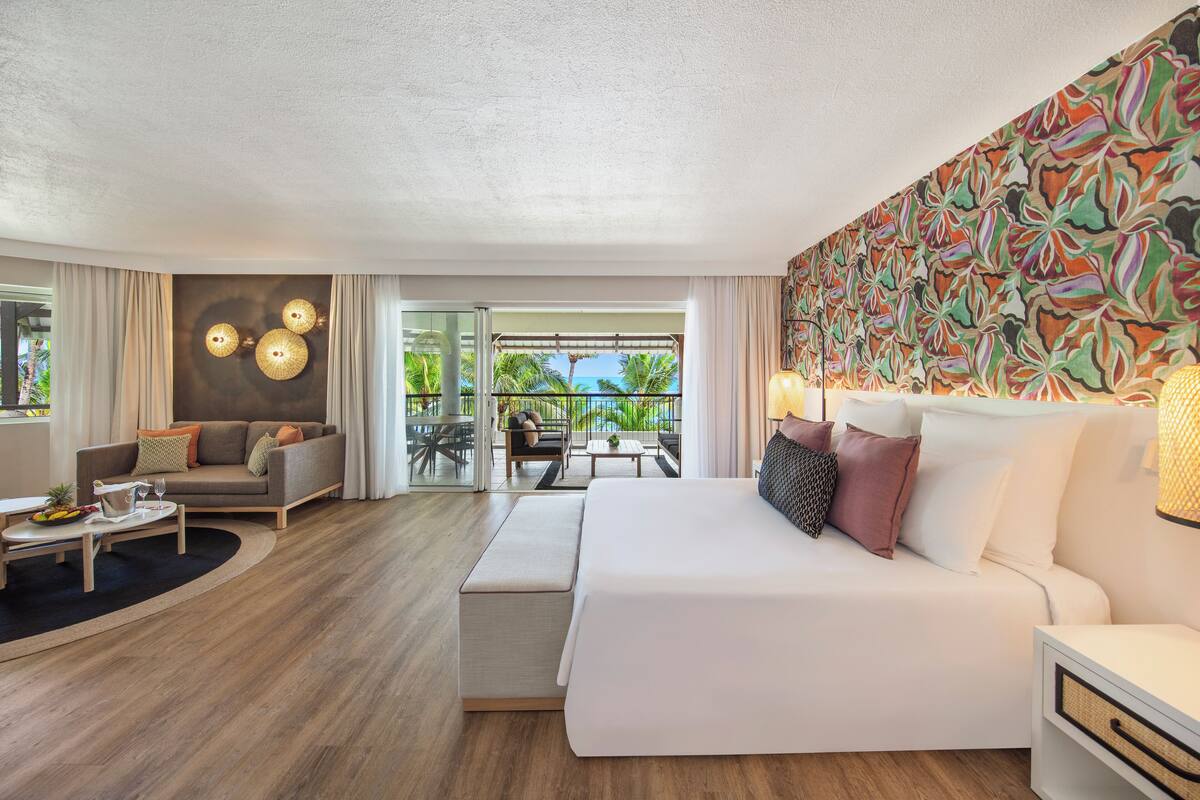 Habitación de hotel moderna con una cama grande, un sofá, zona de comedor y balcón con vistas a palmeras.
