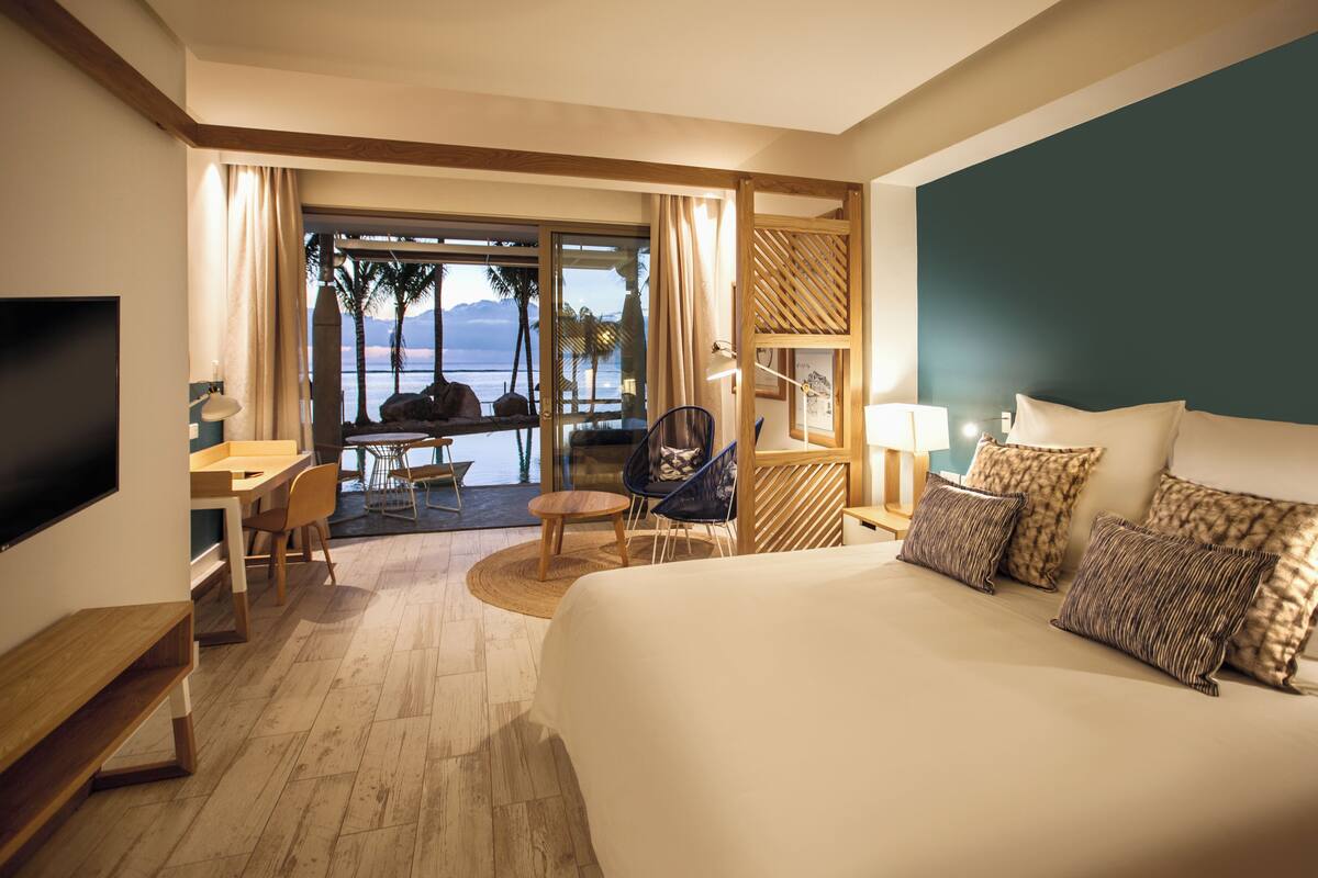 Habitación de hotel con una cama grande, un escritorio con una silla, un televisor y vista al océano a través de una puerta de vidrio.