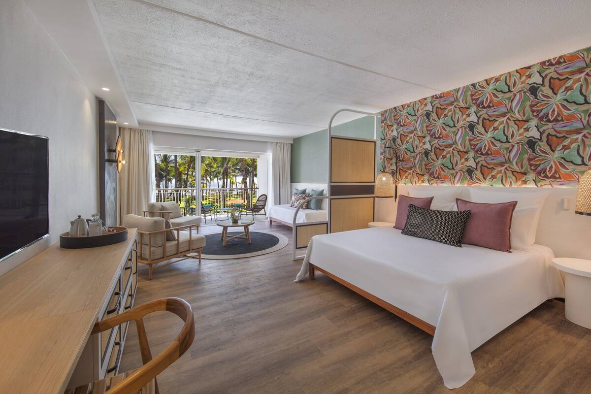 Habitación de hotel moderna con una cama grande, un escritorio, un televisor y un balcón con mobiliario exterior.