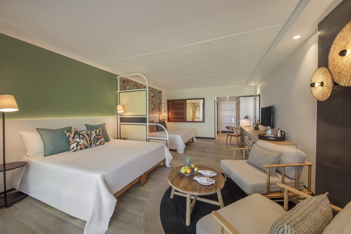 Una habitación de hotel moderna con dos camas, un sofá, una pequeña mesa de comedor con fruta y un televisor.