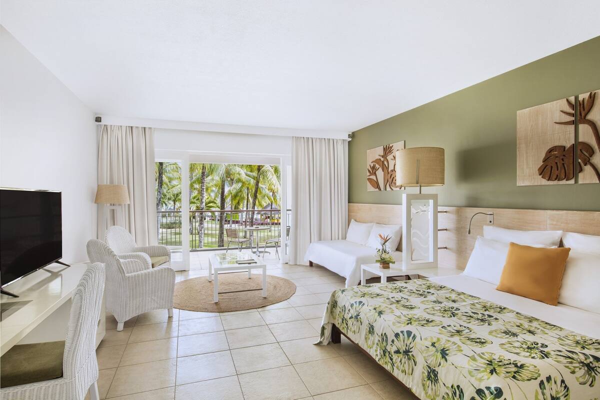 Habitación de hotel con cama, televisor, sofá, mesa de centro y vistas a palmeras.