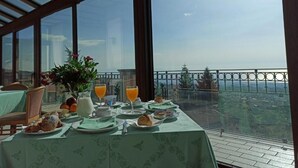 Une table de petit-déjeuner dressée avec des viennoiseries, un panier de fruits, une carafe de lait et un verre de jus d’orange, donnant sur un paysage magnifique à travers de grandes fenêtres.