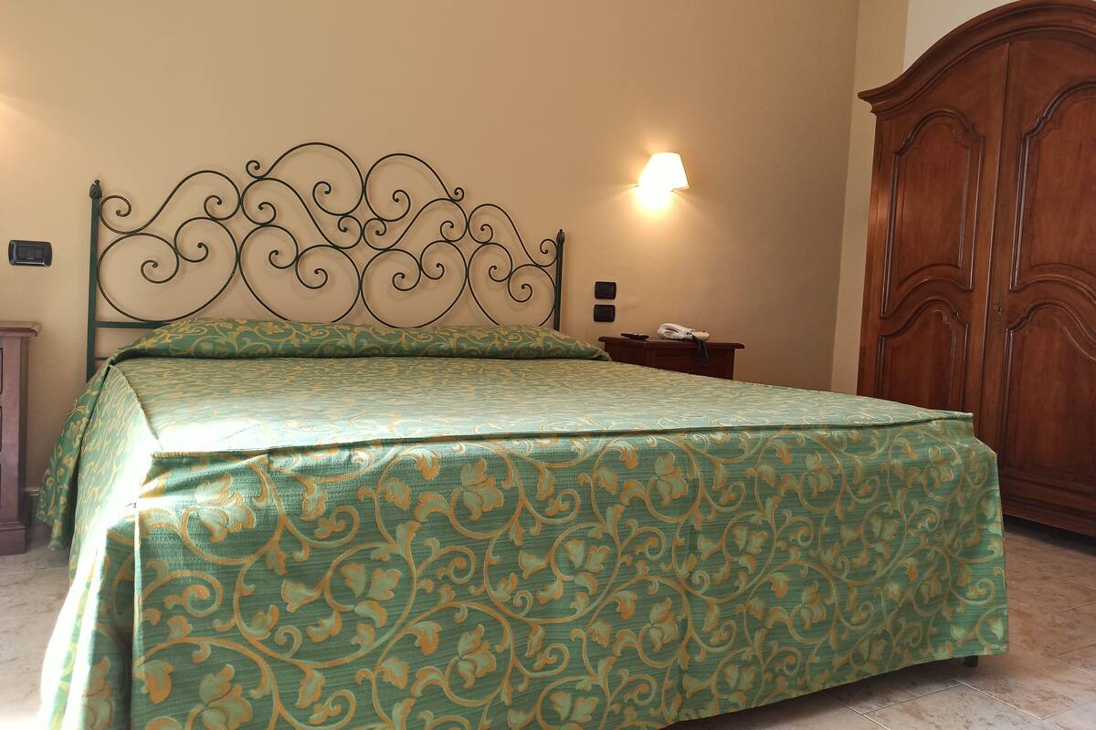 A neatly made bed with a patterned bedspread, a bedside table with a lamp, and a wooden wardrobe in a room with beige walls.