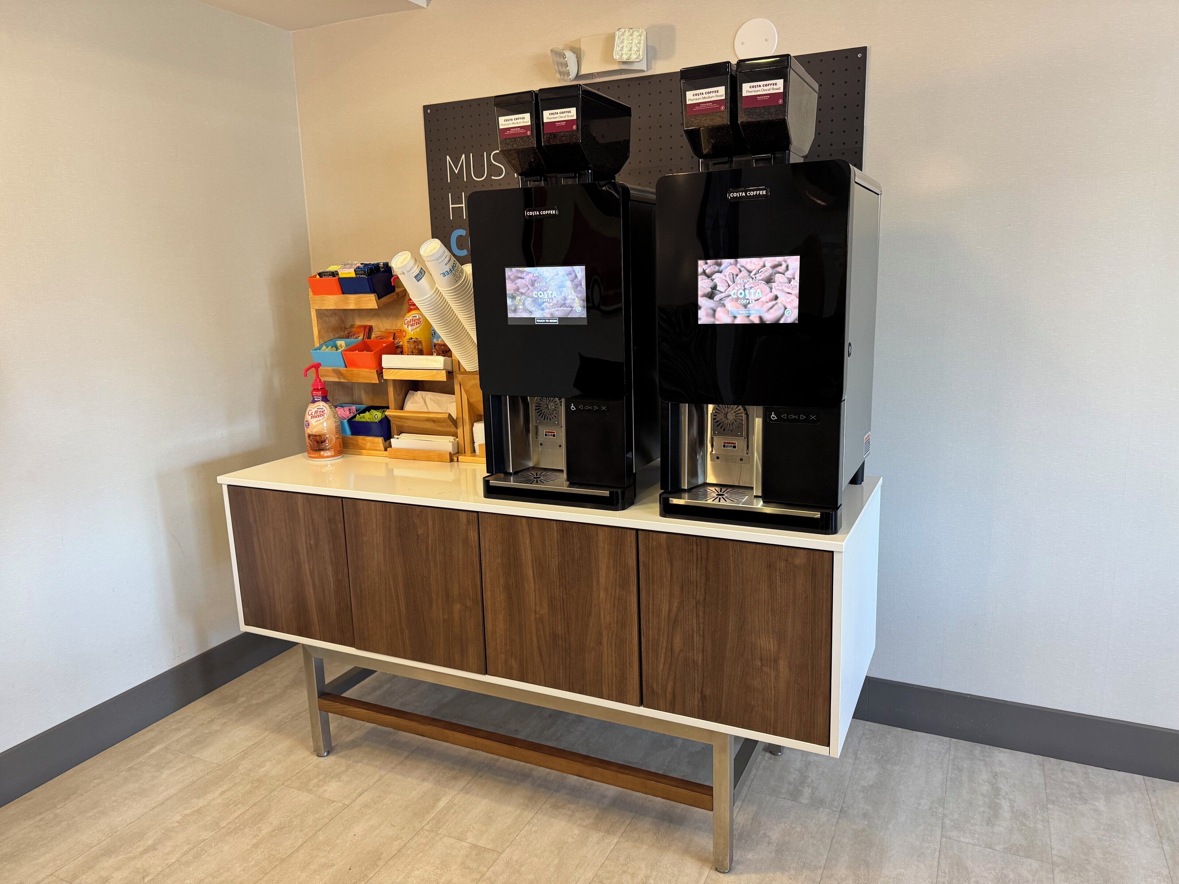 Un espace dédié au café, équipé de deux machines à expresso, d’un distributeur de café et d’une variété de capsules de café, le tout installé sur un comptoir en bois.