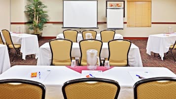 Sala de conferência com mesas, cadeiras, tela de projeção e um vaso de planta.