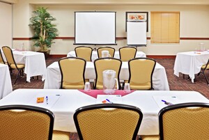 Sala de conferência com mesas, cadeiras, tela de projeção e um vaso de planta.