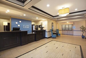 Zona de recepción de hotel con mostrador, pared azul con un logotipo, área de descanso con una silla y un ventanal con cortinas.