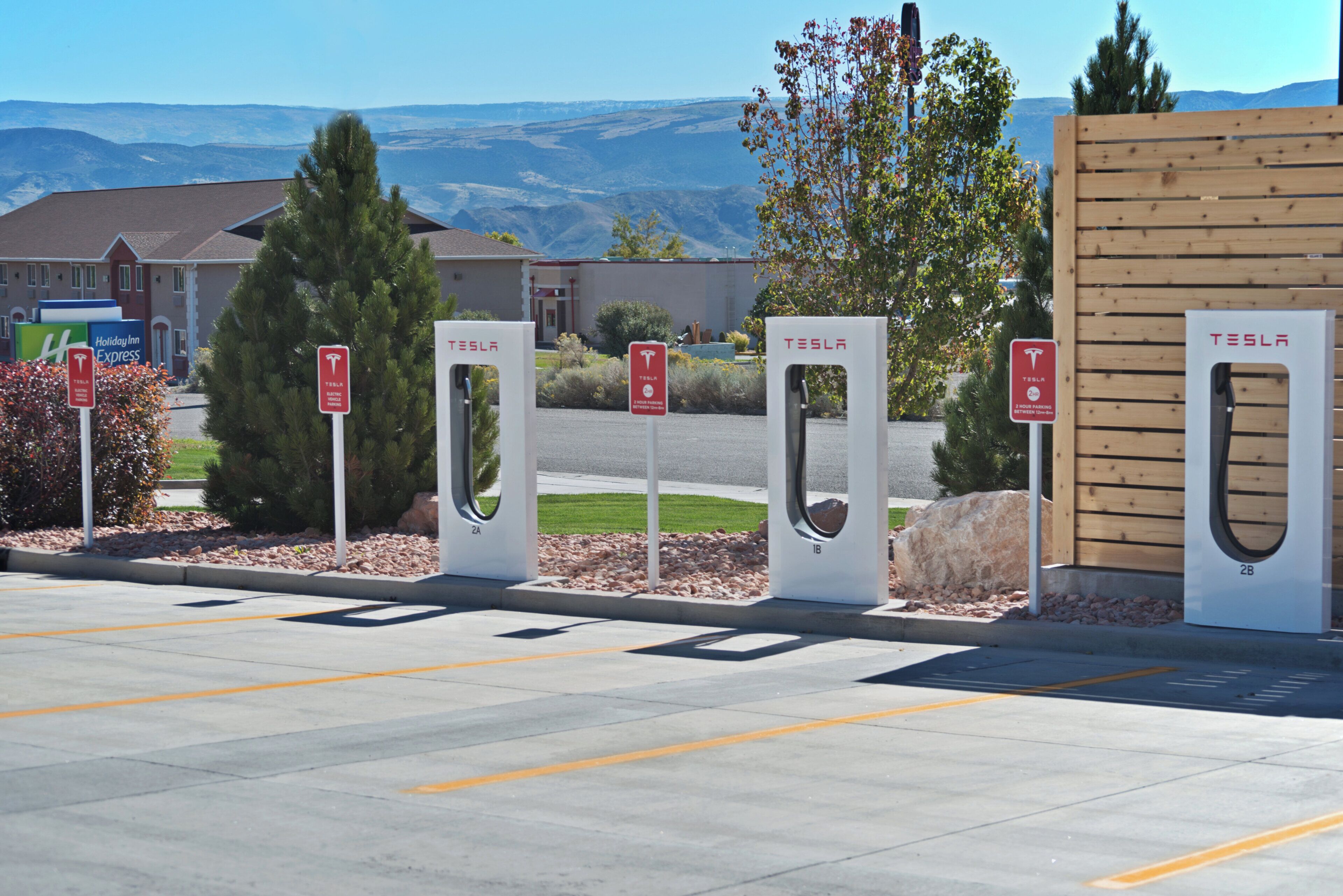 A parking area with multiple Tesla charging stations, a Holiday Inn Express sign, and a building in the background.