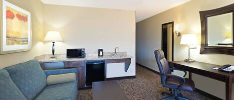 A hotel room with a kitchenette, a desk, a chair, a sofa, and a bed.